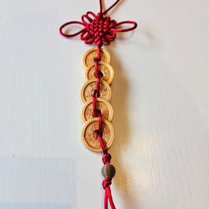 May include: A red Chinese knot with a decorative flower and a loop for hanging. Below the knot are five wooden coins strung together with red string. A small brown bead is at the bottom of the coins.