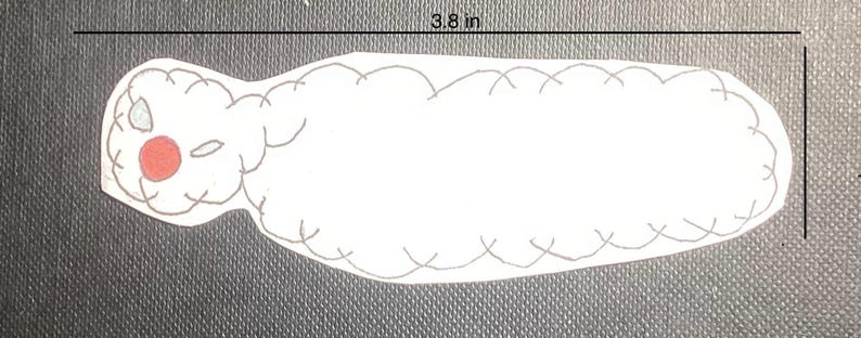 K&ouml;nnte beinhalten: Wei&szlig;er Filz-Applique einer Raupe mit roter Nase und schwarzen Augen. Die Raupe ist 9,7 cm lang.
