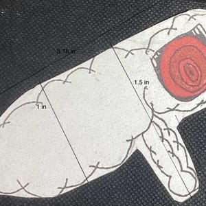 K&ouml;nnte beinhalten: Eine wei&szlig;e Filzapplikation in Form einer Hand mit einem roten, wirbelnden Auge. Die Applikation ist 9,5 cm lang und 3,8 cm breit.