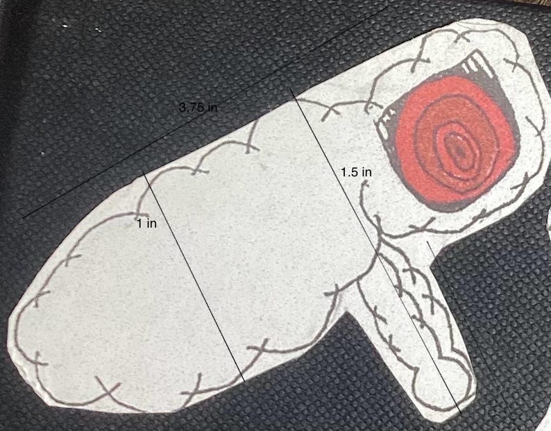 K&ouml;nnte beinhalten: Eine wei&szlig;e Filzapplikation in Form einer Hand mit einem roten, wirbelnden Auge. Die Applikation ist 9,5 cm lang und 3,8 cm breit.