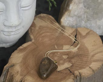 Op de afbeelding: Een gepolijste, donkerbruine, onregelmatig gevormde stenen hanger aan een dunne, goudkleurige metalen ketting. De steen heeft subtiele, lichtere aders over het gladde, glanzende oppervlak, wat zorgt voor een natuurlijke en reflecterende uitstraling.