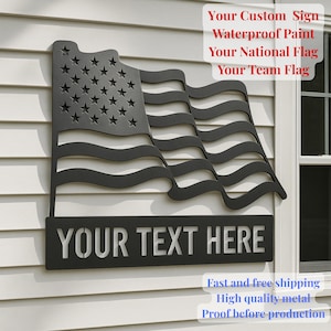 Peut inclure: Panneau en métal noir en forme de drapeau américain flottant. Le panneau présente des étoiles et des rayures découpées, avec le texte "YOUR TEXT HERE" en dessous. Il est monté sur un mur blanc.