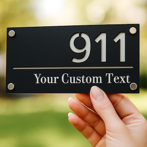 May include: Black rectangular sign with the number "911" cut out in white. The text "Your Custom Text" is below the number. The sign is held by a hand and has silver-colored mounting hardware.
