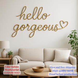 May include: Gold metal wall art spelling "hello gorgeous" in cursive script. The word "gorgeous" ends with a heart shape. The art is displayed above a beige sofa in a living room setting.