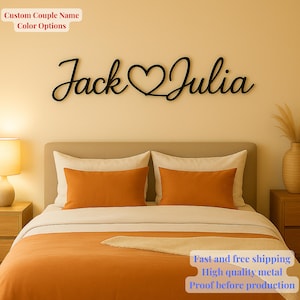 May include: Black metal wall art with the names "Jack" and "Julia" in a cursive font, joined by a heart. The artwork hangs above a bed with orange bedding and white pillows. Text at the top says "Custom Couple Name Color Options".