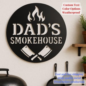 May include: Black metal sign with the words "DAD'S SMOKEHOUSE" cut out, featuring flames and crossed cleavers. The sign is round and has the text "Custom Text Color Options Weatherproof".