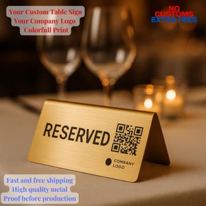 May include: A gold-colored metal table sign with the word "RESERVED" in black. A QR code and a company logo are also printed on the sign. The sign is angled and stands on a table. The background includes wine glasses and candles.