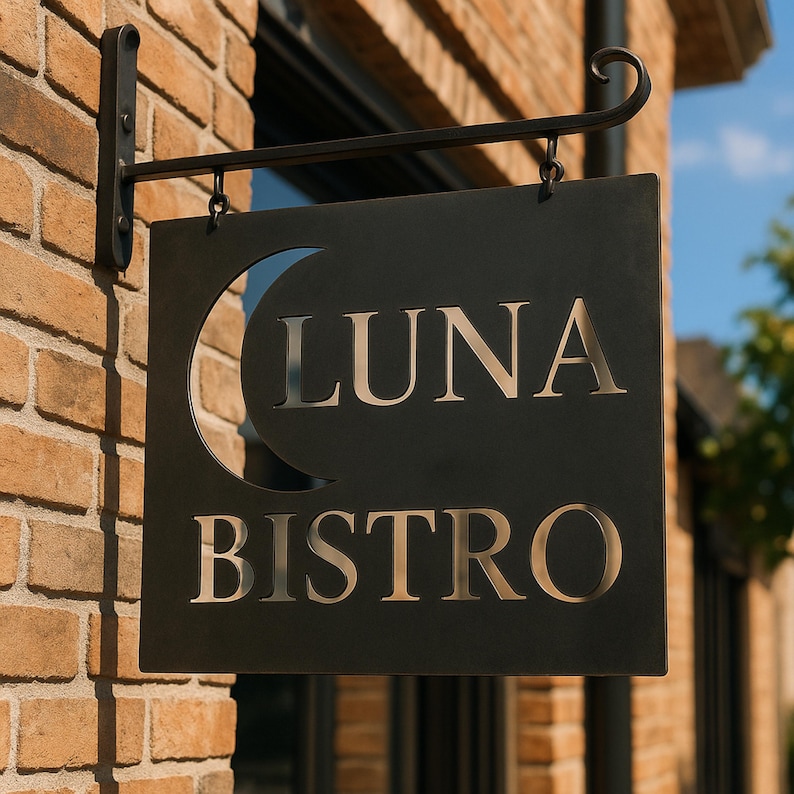 May include: A black metal sign for "LUNA BISTRO" with a crescent moon cutout. The sign is square and hangs from a decorative black metal bracket attached to a brick wall. The letters are cut out, revealing the background.