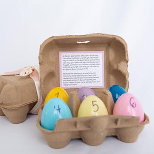 Könnte beinhalten: Ein Oster-Bastelset mit einem Eierkarton aus Pappe, der sechs nummerierte Eier enthält. Die Eier sind gelb, blau, rosa und lila und mit den Zahlen 1 bis 6 versehen. Ein zweiter Eierkarton ist mit einem rosa Band gebunden.