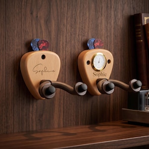 May include: Two light-coloured wooden guitar wall hangers with dark metal arms. Each hanger features a guitar pick holder at the top and the name Sophie engraved on the front. The right hanger includes a small round gold-rimmed analogue hygrometer.
