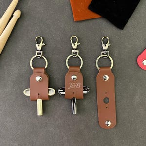 Pode incluir: Três porta-chaves de couro castanho com ferragens de metal e fechos de mosquetão. Dois porta-chaves seguram chaves de afinação de bateria, uma com acabamento dourado e outra prateado. O porta-chaves central apresenta as letras gravadas J&B.