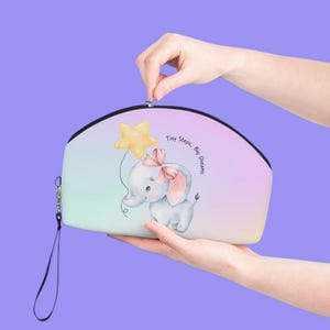 May include: A light blue and pink zippered pouch with a watercolor illustration of a baby elephant holding a yellow star balloon. The pouch has a black zipper and a black strap. The text "Tiny Steps, Big Dreams" is printed on the pouch.