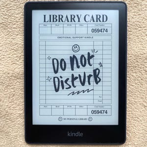 May include: A black Kindle e-reader displaying a custom "Library Card" design. The card reads "EMOTIONAL SUPPORT KINDLE" and "Do Not Disturb" in handwritten style. The Kindle is on a beige surface.