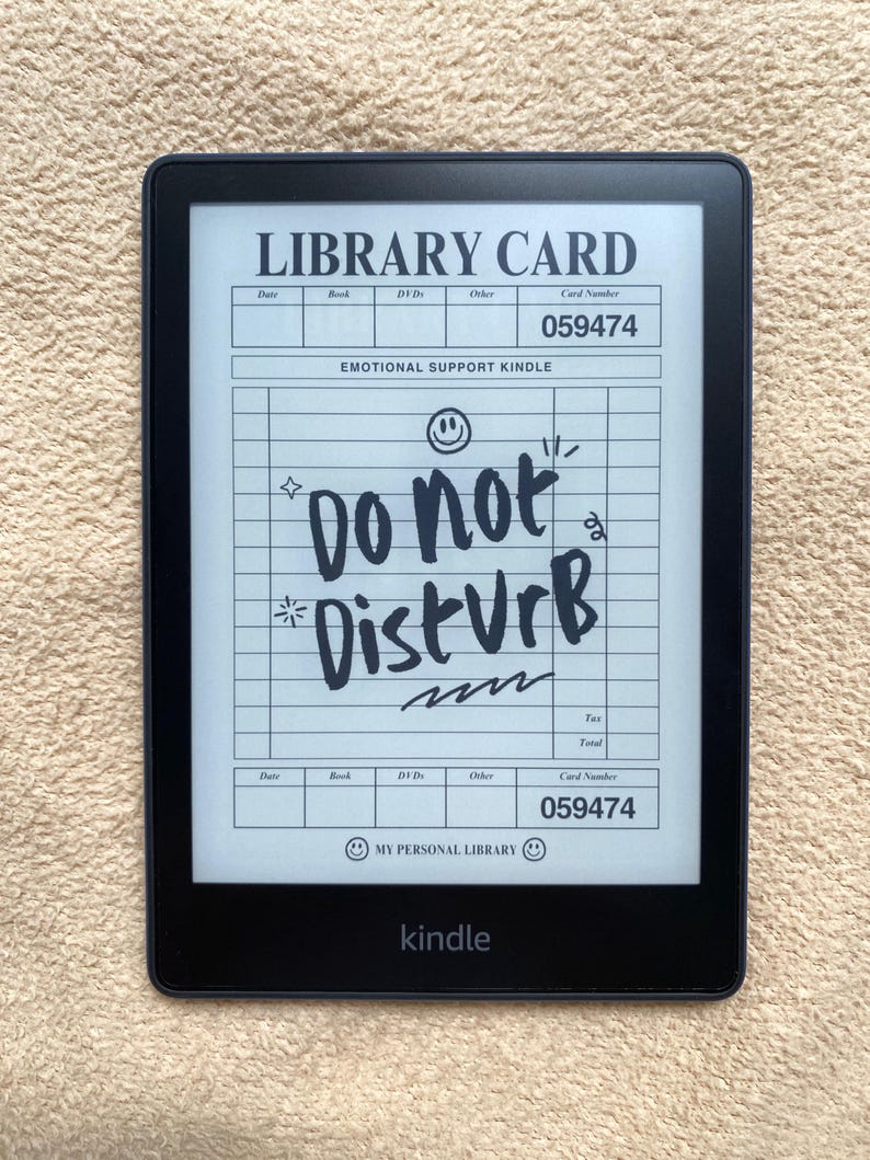 May include: A black Kindle e-reader displaying a custom "Library Card" design. The card reads "EMOTIONAL SUPPORT KINDLE" and "Do Not Disturb" in handwritten style. The Kindle is on a beige surface.