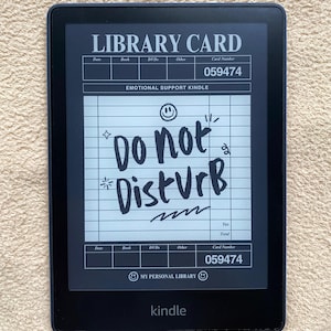 May include: A black Kindle e-reader displaying a custom "Library Card" screen. The screen reads "Do Not Disturb" in handwritten style, with a card number of 059474. The device is on a beige surface.