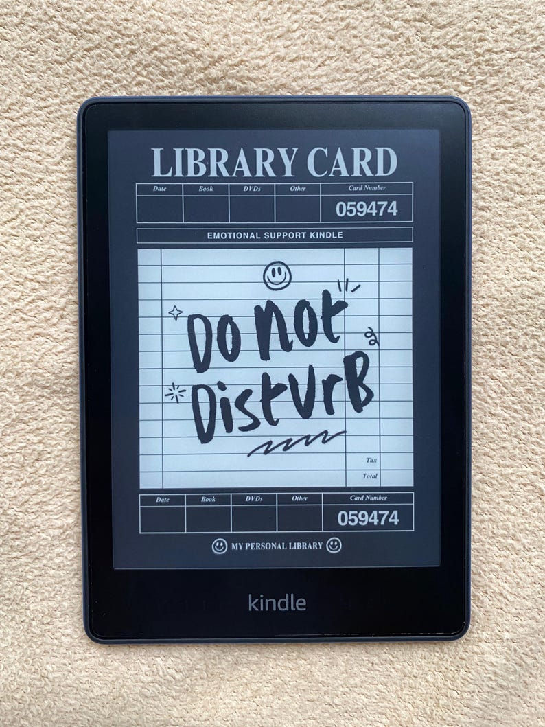 May include: A black Kindle e-reader displaying a custom "Library Card" screen. The screen reads "Do Not Disturb" in handwritten style, with a card number of 059474. The device is on a beige surface.