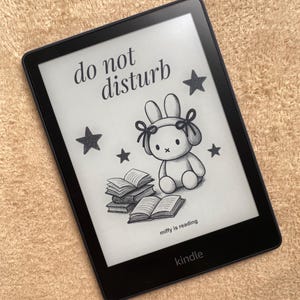 Puede incluir: Lector de libros electrónicos Kindle negro que muestra un boceto de un conejito con lazos, estrellas y libros. El texto en la pantalla dice "do not disturb" y "miffy is reading". El lector está sobre una superficie beige.