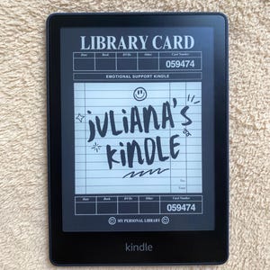 May include: A black Kindle e-reader displaying a custom library card design. The screen reads "LIBRARY CARD" at the top, with "JULIANA'S KINDLE" written in a handwritten style. The device also shows the word "Kindle" at the bottom.