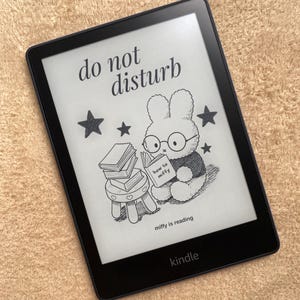 May include: A black Kindle e-reader displaying a cartoon illustration of a bunny reading a book with the text "do not disturb". The image also includes the text "miffy is reading" and the Kindle logo.