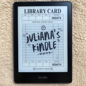 May include: A black Kindle e-reader displaying a handwritten "Library Card" design with the text "JULIANA'S KINDLE." The screen also shows the words "EMOTIONAL SUPPORT KINDLE" and "MY PERSONAL LIBRARY."