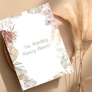 Könnte beinhalten: Ein weißer, druckbarer Planer mit einem floralen Design mit beigefarbenen, braunen und rosa Blüten und Blättern. Der Text "The Monthly Money Planner" ist in Grau auf der Vorderseite des Planers geschrieben.
