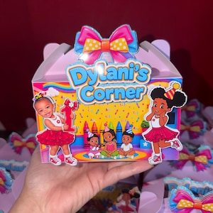 May include: A light purple party favor box featuring a large pink and yellow polka dot bow on top. The front displays the text "Dylani's Corner" in blue, surrounded by colorful illustrations of children, crayons, and a rainbow on a yellow background.