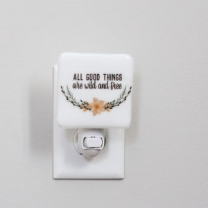 Op de afbeelding: Witte keramische nachtlamp met een bloemenmotief en de tekst "All good things are wild and free".