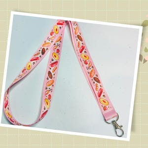 May include: A pink lanyard with a repeating pattern of baked goods such as donuts, bread, and pastries. It has a silver-coloured clasp for attaching keys or an ID badge. The design is colourful and playful, suitable for everyday use.
