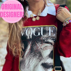 May include: Red sweatshirt with a white collar featuring a Vogue Christmas Edition cover. The cover art is a black and white image of a person. A gold necklace and bracelets are also visible.