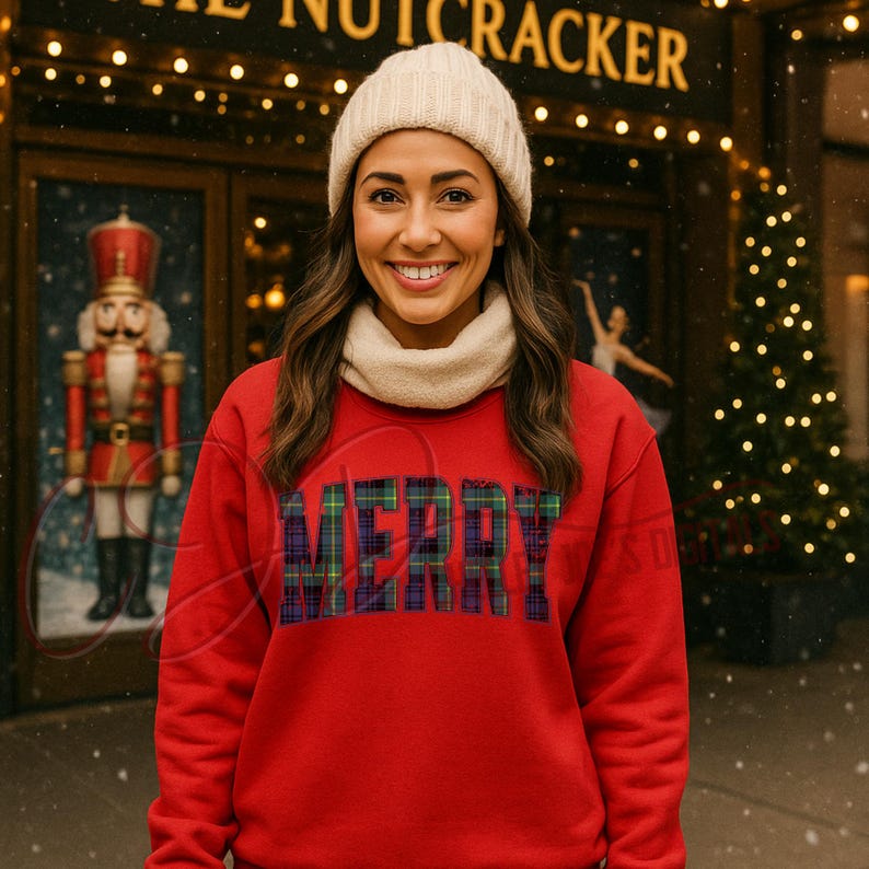 Op de afbeelding: Een rode sweatshirt met het woord "MERRY" in een geruit patroon. De persoon op de foto draagt een cr&egrave;mekleurige muts en sjaal. De achtergrond bevat een gebouw met de woorden "THE NUTCRACKER" en een kerstboom.