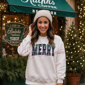 Op de afbeelding: Een cr&egrave;mekleurige sweatshirt met het woord "MERRY" in een ruitpatroon. De persoon op de foto draagt een beige muts. De achtergrond omvat een koffiehuis en kerstversieringen.
