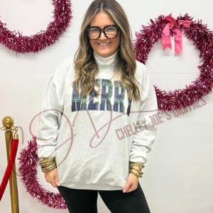 Op de afbeelding: Een lichtgrijs sweatshirt met het woord "MERRY" in een blauw en groen geruit ontwerp. De persoon draagt een zwarte bril en gouden armbanden. Twee roze slingerskransen zijn op de achtergrond.