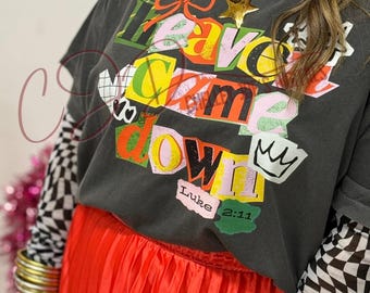 Op de afbeelding: Donkergrijs T-shirt met de woorden "Leave, Came, Down" in een kleurrijk blokletterontwerp. Het shirt heeft een ronde hals en korte mouwen. De tekst "Luke 2:11" is onderaan gedrukt. Het shirt wordt gecombineerd met een rode geplooide rok.