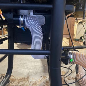 May include: Close-up of a black table saw frame with a clear, flexible hose attached. The hose is connected to a dust collection system, with a blue clamp visible. The workshop setting includes other tools.
