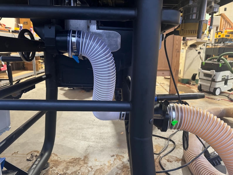 May include: Close-up of a black table saw frame with a clear, flexible hose attached. The hose is connected to a dust collection system, with a blue clamp visible. The workshop setting includes other tools.