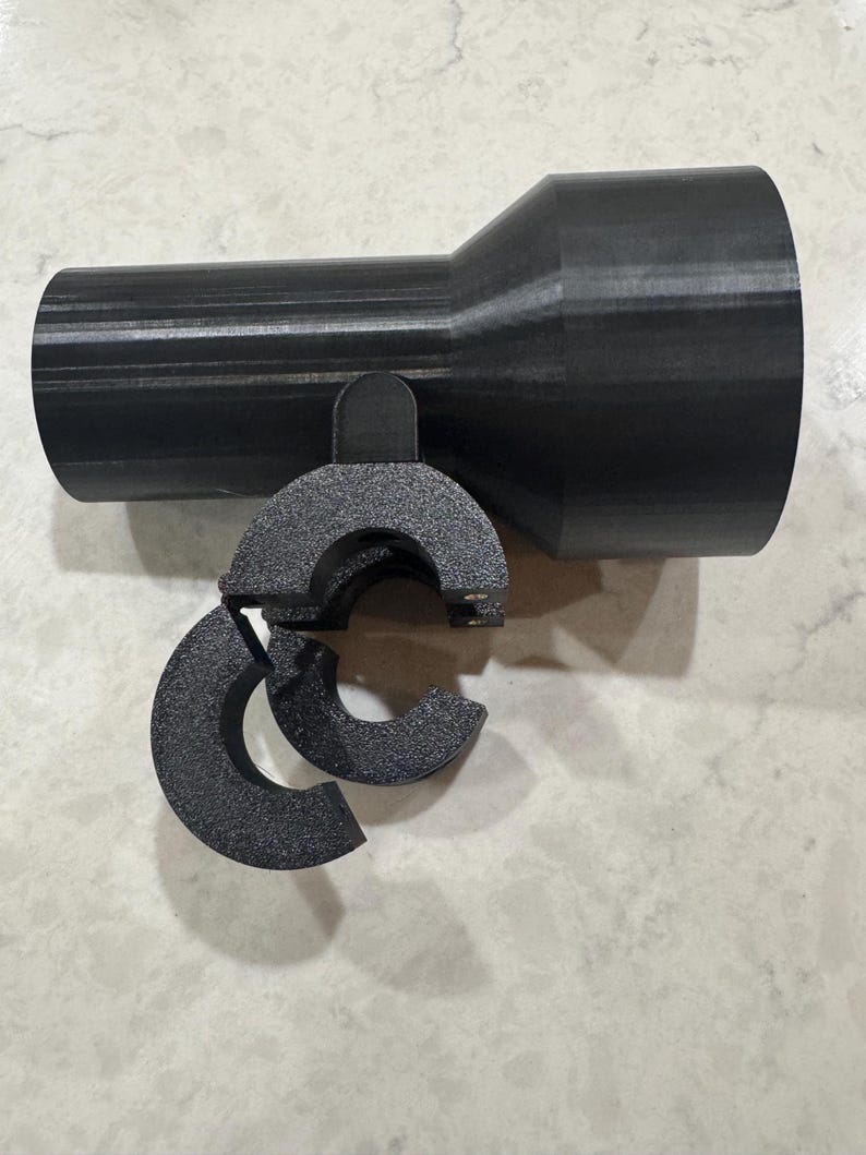 May include: A black plastic pipe fitting with a cylindrical body and a flared end. A curved, split clamp is attached to the body. The object is designed for connecting pipes or hoses.