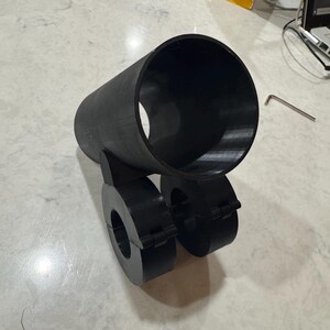 May include: A black, cylindrical object with two circular components attached. The object appears to be 3D-printed and sits on a light-colored surface. A small metal tool is visible in the background.