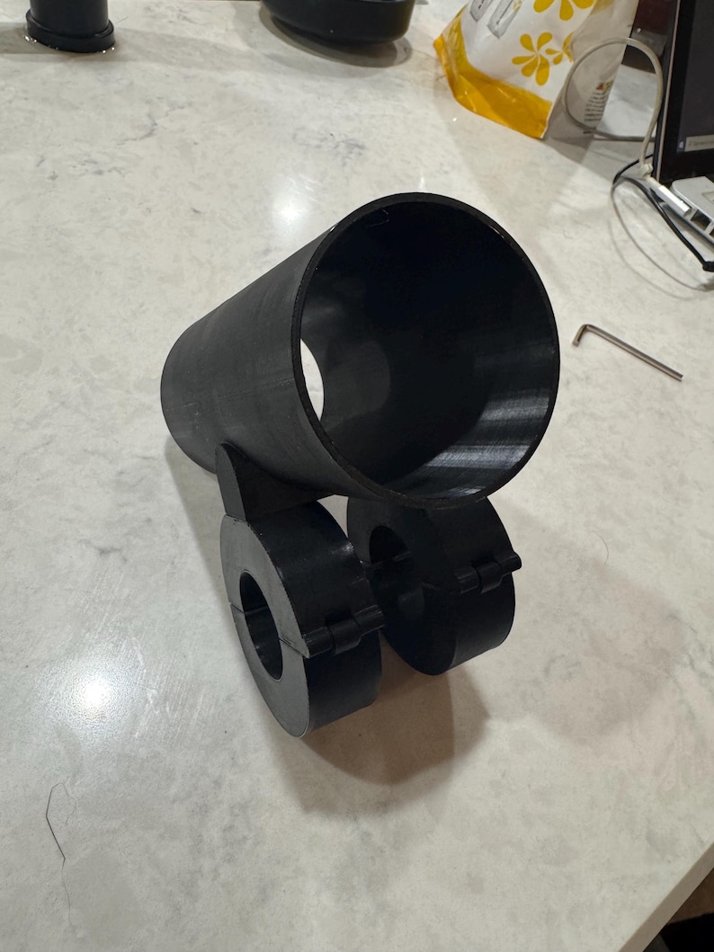 May include: A black, cylindrical object with two circular components attached. The object appears to be 3D-printed and sits on a light-colored surface. A small metal tool is visible in the background.