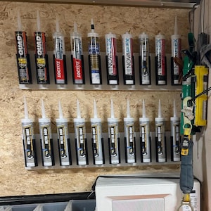 May include: A selection of caulk tubes, including brands such as Quad, Loctite, and NovaFlex, are arranged on a wall-mounted rack. The tubes are in various colours and sizes, with white nozzles and black labels. The image shows a variety of construction and sealant products.