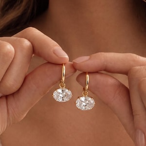Puede incluir: Un par de pendientes de aro dorados con piedras transparentes facetadas de forma ovalada suspendidas de cada aro. Los pendientes tienen un acabado dorado pulido y las piedras transparentes están engastadas en un marco de metal simple.