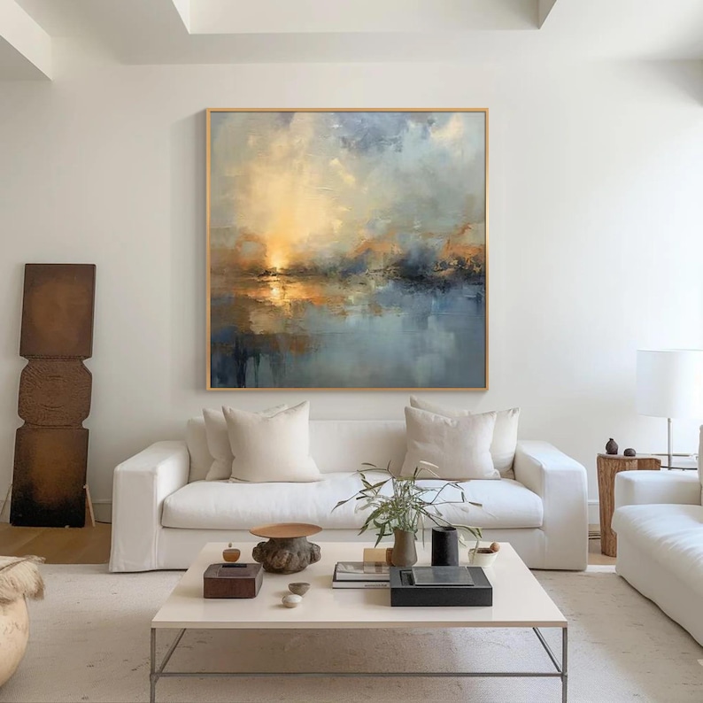 Puede incluir: Gran pintura abstracta de paisaje con marco dorado. La obra presenta un atardecer sobre el agua, con tonos azules, amarillos y naranjas. La pintura cuelga sobre un sof&aacute; blanco en un sal&oacute;n moderno.