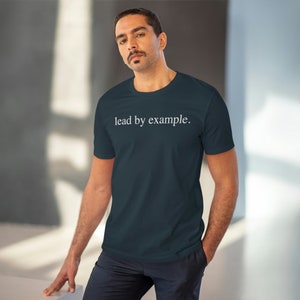 Peut inclure: Un t-shirt bleu marine avec l'inscription blanche "lead by example." centrée sur la poitrine. Le t-shirt est de style col rond et semble être fait d'une matière douce. La personne porte le t-shirt.