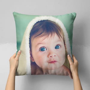 Puede incluir: Un cojín decorativo con la cara de un bebé con ojos azul brillante y una toalla con capucha blanca y suave. El cojín tiene un fondo verde claro y está sostenido por dos manos.