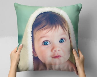 Può includere: Un cuscino decorativo con il volto di un bambino con occhi azzurri brillanti e un morbido asciugamano con cappuccio bianco. Il cuscino ha uno sfondo verde chiaro ed è tenuto da due mani.