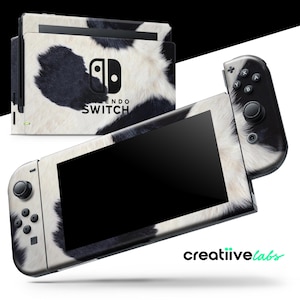 Op de afbeelding: Een Nintendo Switch-console en dock met een zwart-wit koeienhuidpatroon. De console heeft een zwart scherm en zwarte controllers. De tekst "NINTENDO SWITCH" is zichtbaar.
