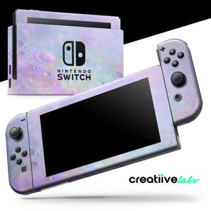 Puede incluir: Una consola Nintendo Switch y mandos con una piel nacarada con temática de galaxia. La consola presenta el logotipo de Nintendo Switch. Los mandos tienen botones y joysticks negros. La marca "creatiivelabs" es visible en la esquina inferior derecha.