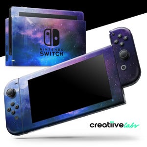 Puede incluir: Una consola Nintendo Switch y sus mandos con un diseño de galaxia. La consola y los mandos están cubiertos con un patrón de nebulosa azul, morado y blanco. El logotipo de Nintendo Switch es visible en la consola.