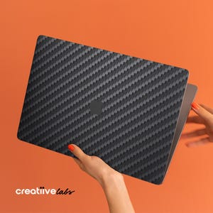 May include: A black laptop with a carbon fiber texture is held against an orange background. The laptop is open, revealing the screen. The brand name "creatiivelabs" is visible in white text at the bottom left.