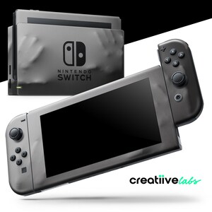 Könnte beinhalten: Eine graue Nintendo Switch-Konsole mit schwarzem Bildschirm und angeschlossenem Controller. Die Konsole wird mit ihrer Dockingstation gezeigt, und der Text "NINTENDO SWITCH" ist auf der Dockingstation zu sehen. Das Logo von "creatiivelabs" befindet sich unten rechts.