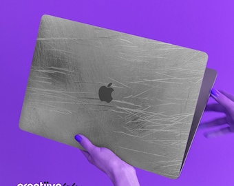 May include: A silver laptop computer with a scratched surface and the Apple logo on the lid. The laptop is being held by a hand with blue nail polish.